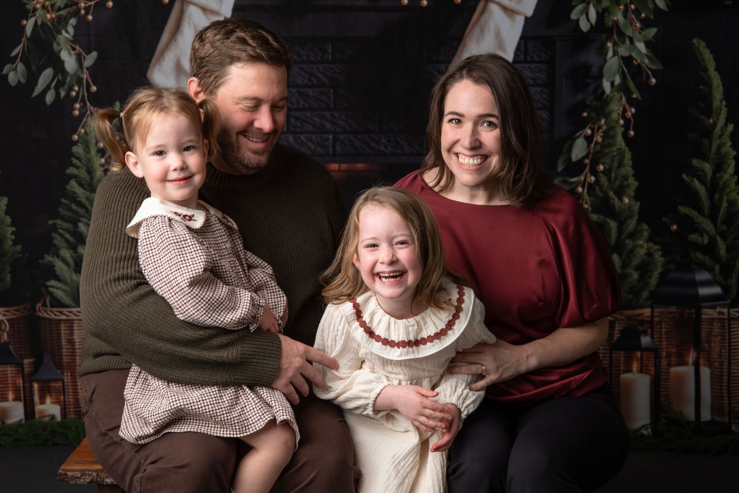A family group poses together for holiday portraits in a cozy indoor setting with greenery and warm lighting.