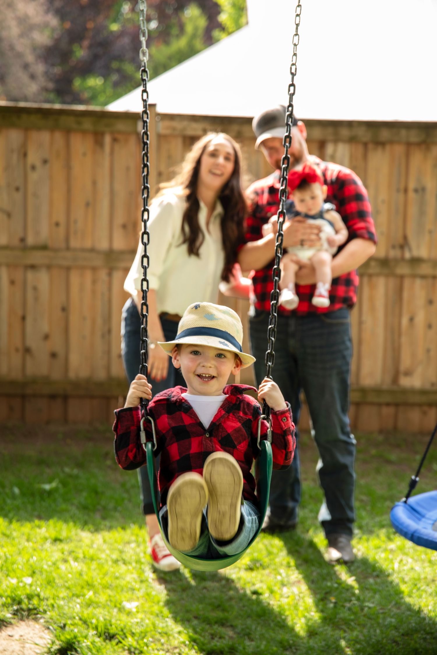 Family enjoying backyard swing set, young boy in plaid shirt and hat swinging while parents stand behind holding baby.