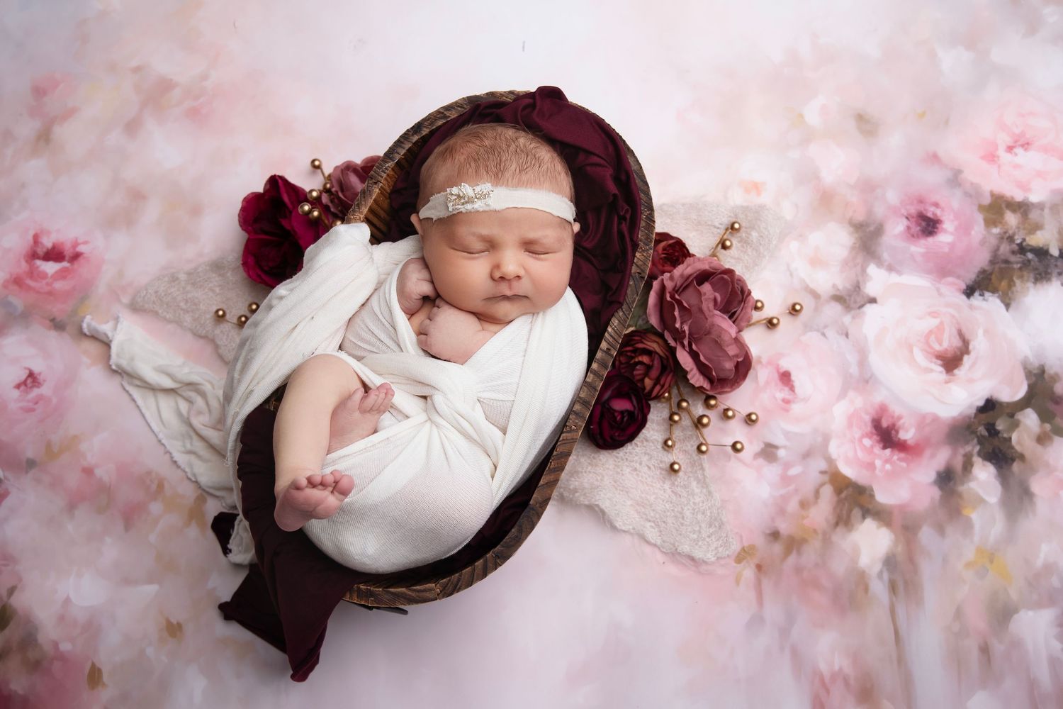 Newborn baby sleeping in dark basket surrounded by pink roses and soft floral backdrop.
