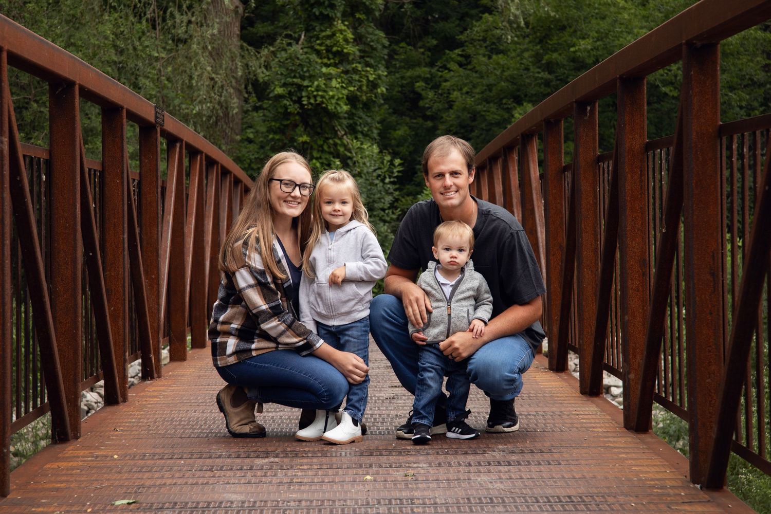 Family poses together on rustic wooden bridge surrounded by lush green forest.