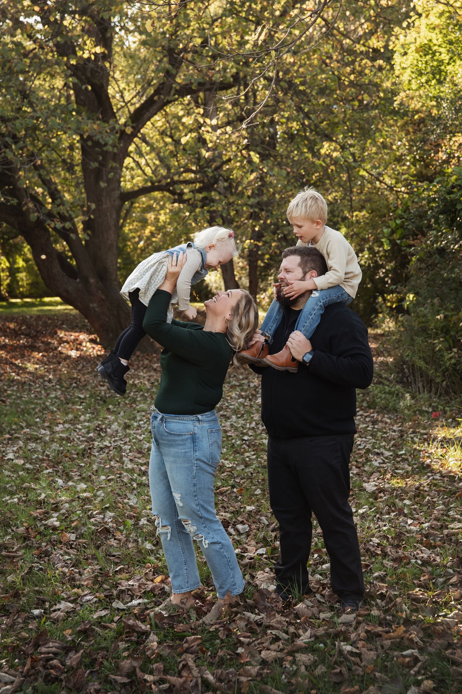 Parents lifting and playing with children under autumn tree in park setting.