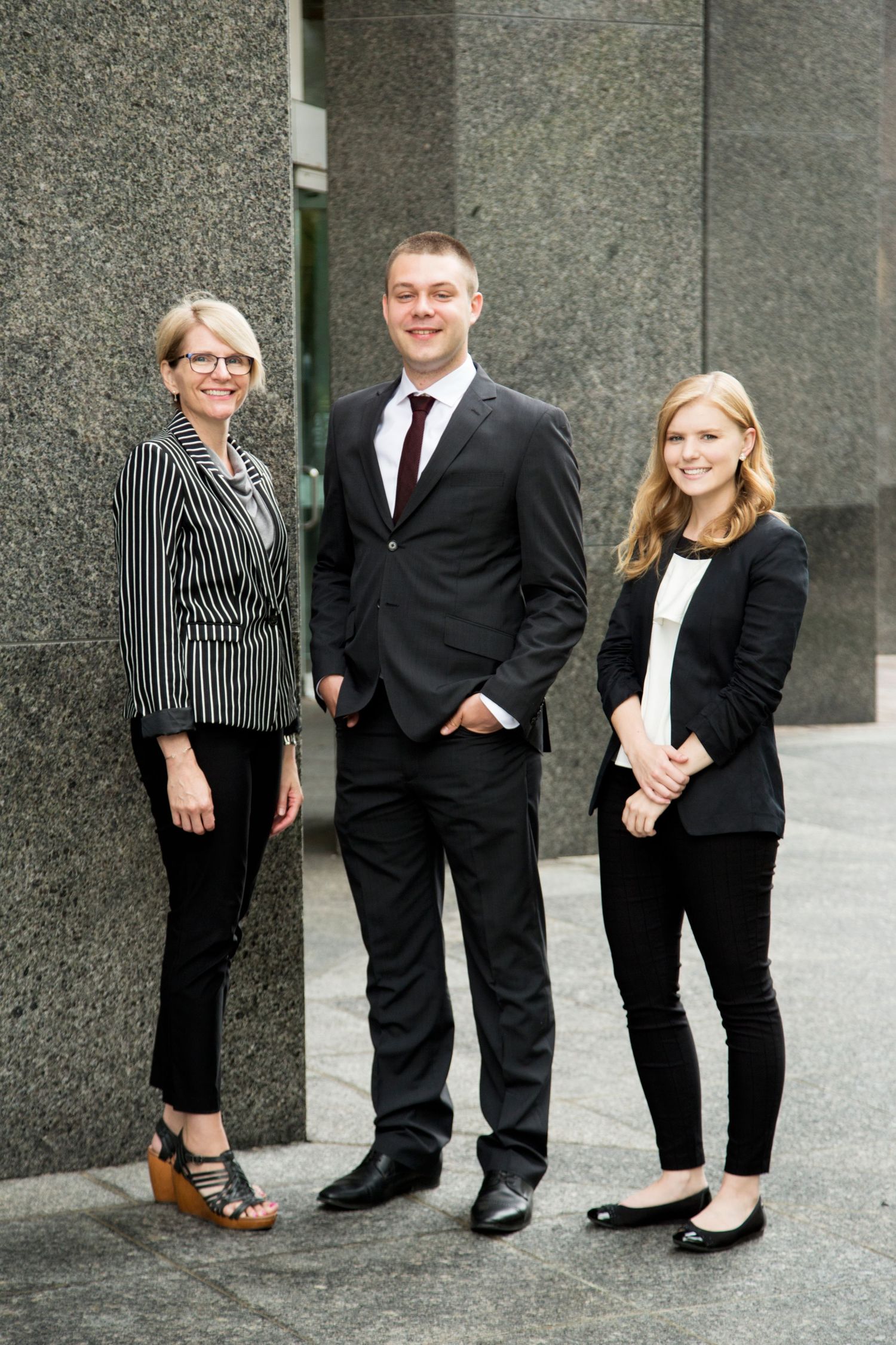 Three business professionals in formal attire pose together against a gray concrete wall.