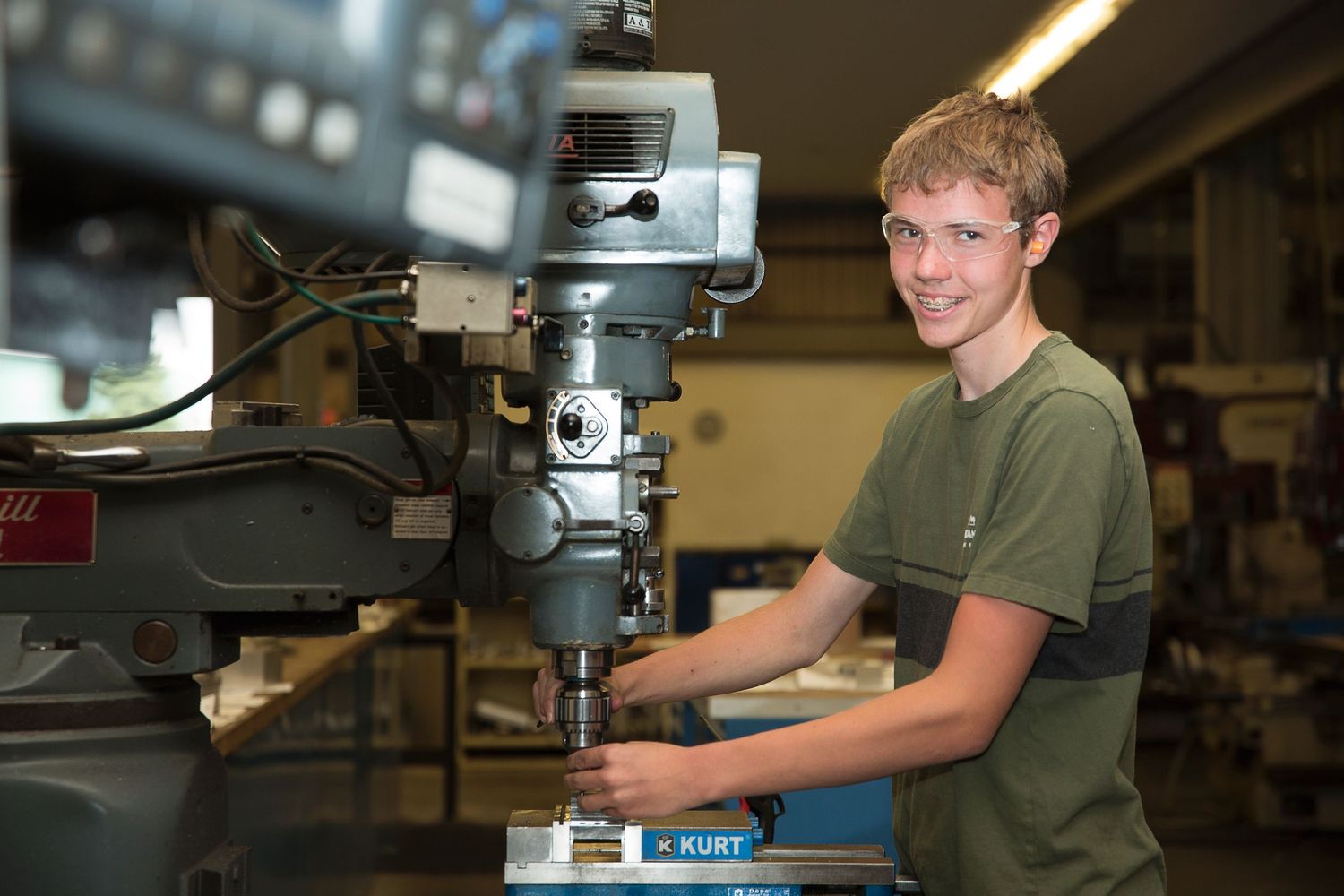 Male student operates a drill press machine at a workstation in a shop classroom while wearing safety gear.