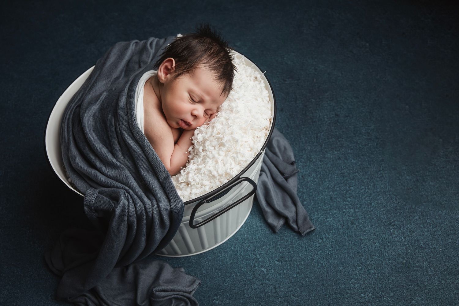 Newborn sleeping peacefully in grey fabric wrap on dark background.