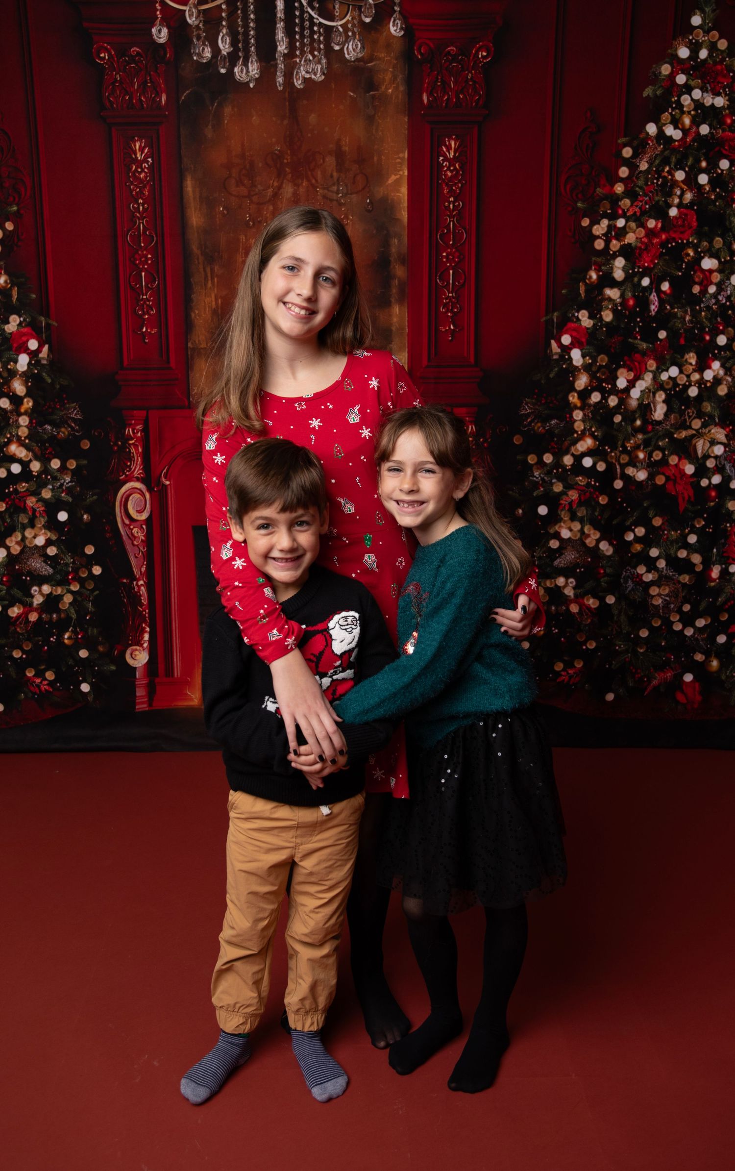 Three children pose together in front of a decorated Christmas backdrop with red walls and twinkling lights.