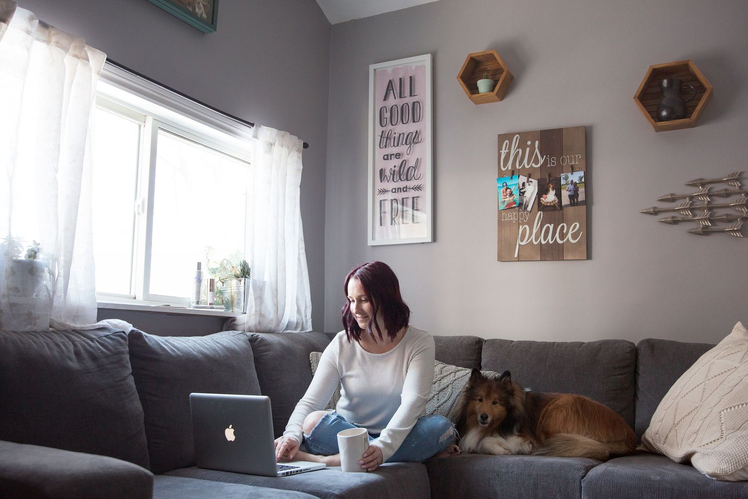 Working from home on laptop computer on gray couch with dog while surrounded by decorative wall art.