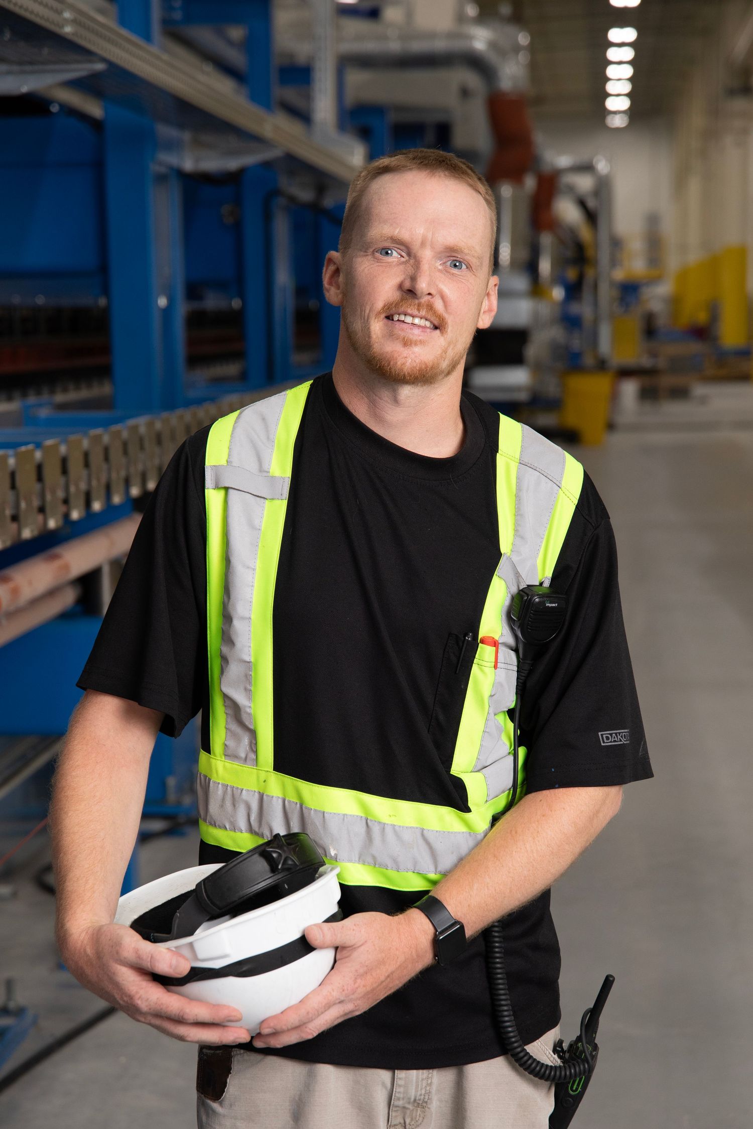 Worker in safety vest and black t-shirt holds protective gear while working in warehouse facility.