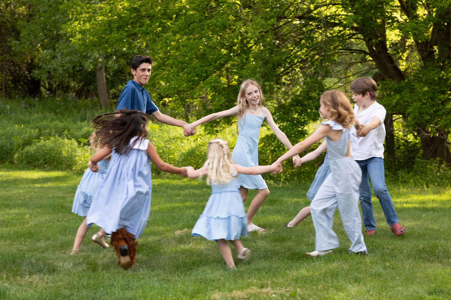Children in blue dresses playing ring-around-the-rosy on green grass surrounded by trees.