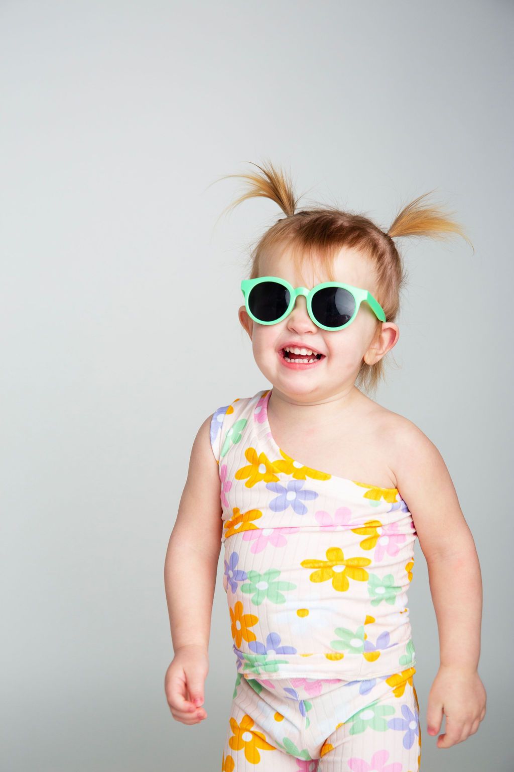 Toddler wearing colorful floral romper and mint sunglasses posing against grey background.