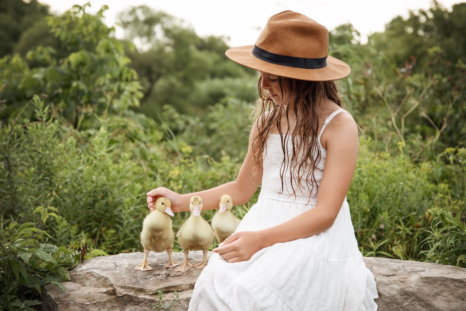 Person in a white summer dress and brown hat sits outdoors with baby chicks in a garden setting.