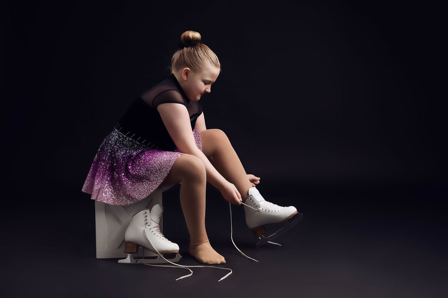 A girl in a sparkly purple dance costume adjusts their ice skate in dramatic lighting.