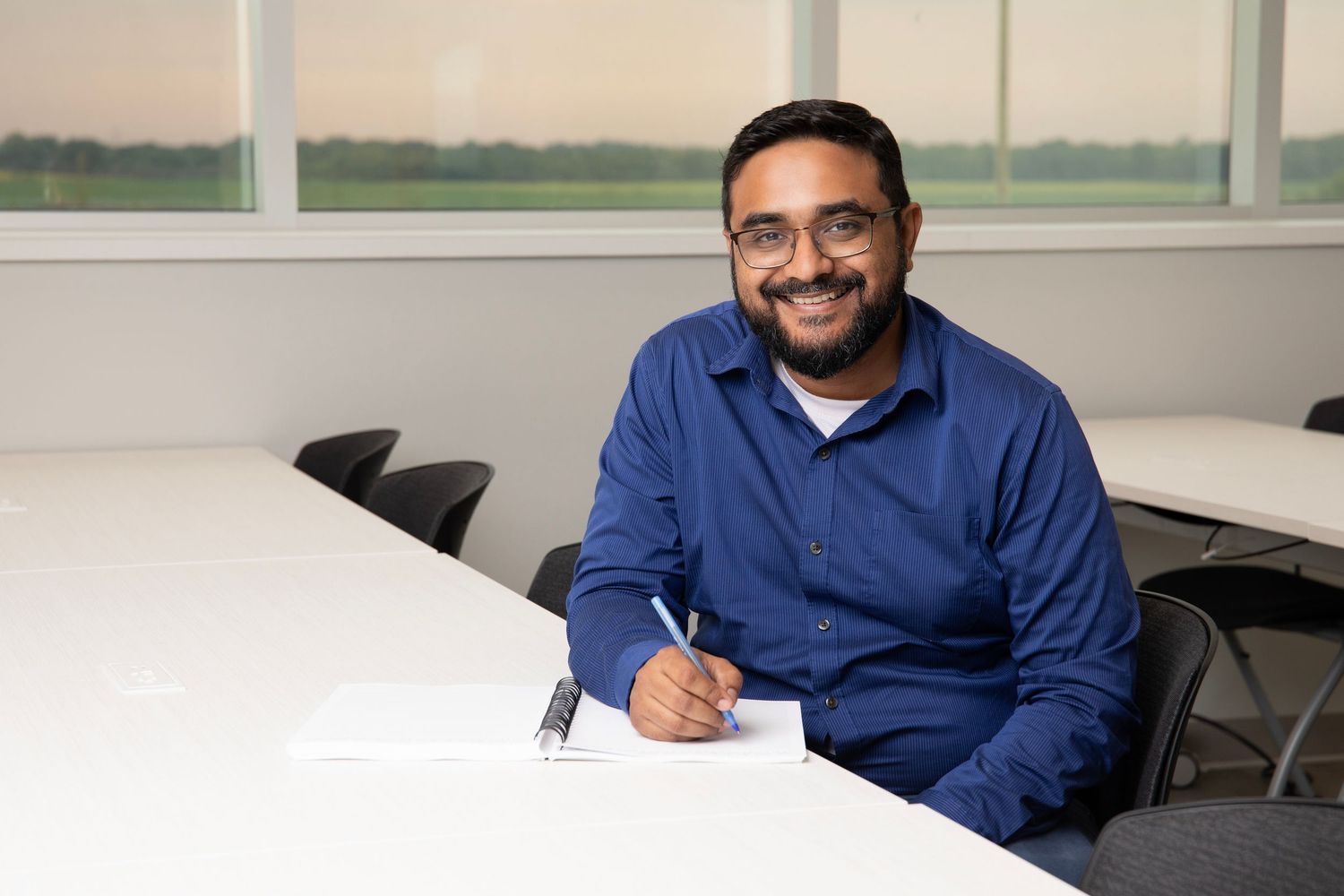 Professional in a blue long-sleeve shirt sits at a white table while writing, looking up with a warm smile.