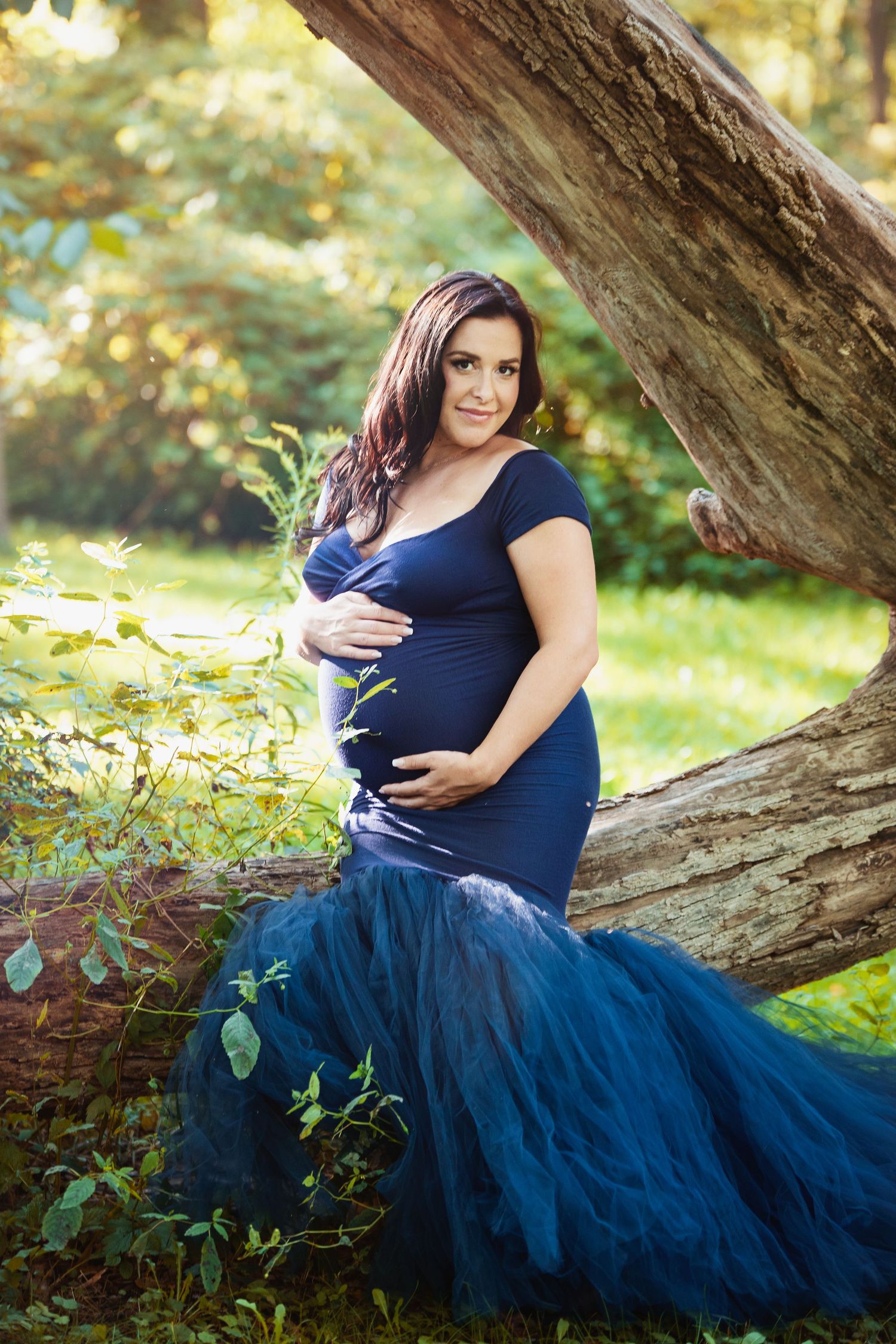 Pregnant woman in navy blue mermaid gown poses elegantly against fallen tree trunk in outdoor maternity shoot.