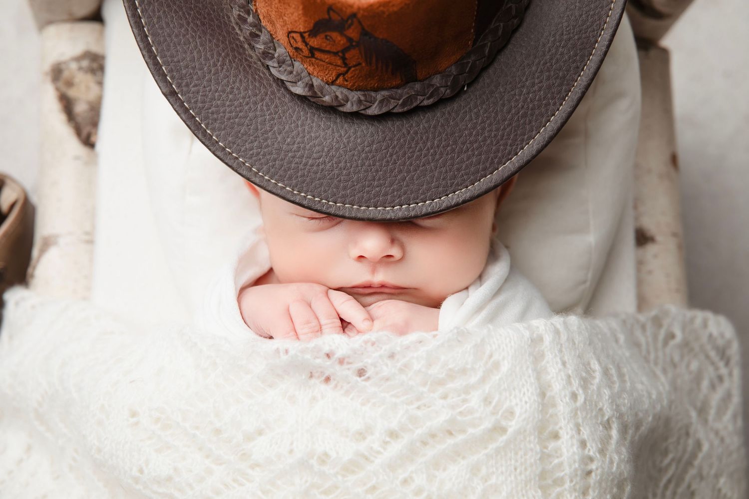 Newborn baby sleeping under brown leather cowboy hat wrapped in white blanket.