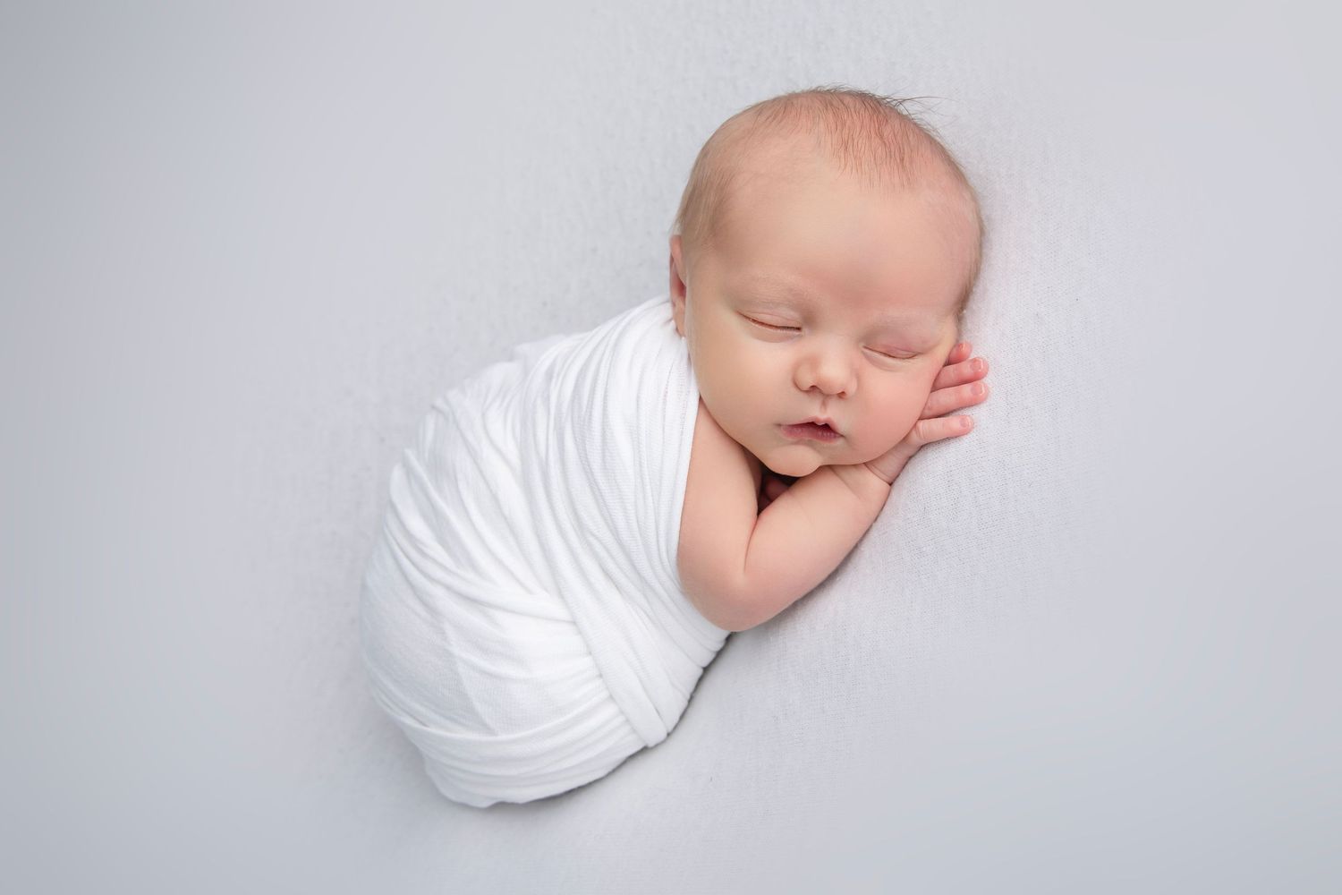Sleeping newborn baby swaddled in white fabric against light background.