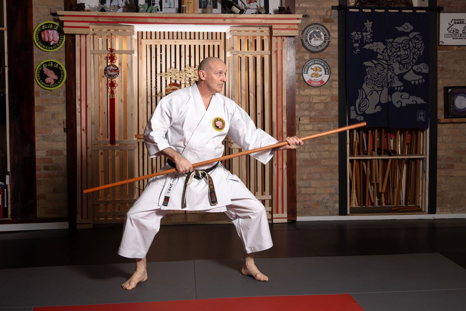 Martial arts instructor in white gi performs bo staff kata stance in a traditional dojo with wooden decor.