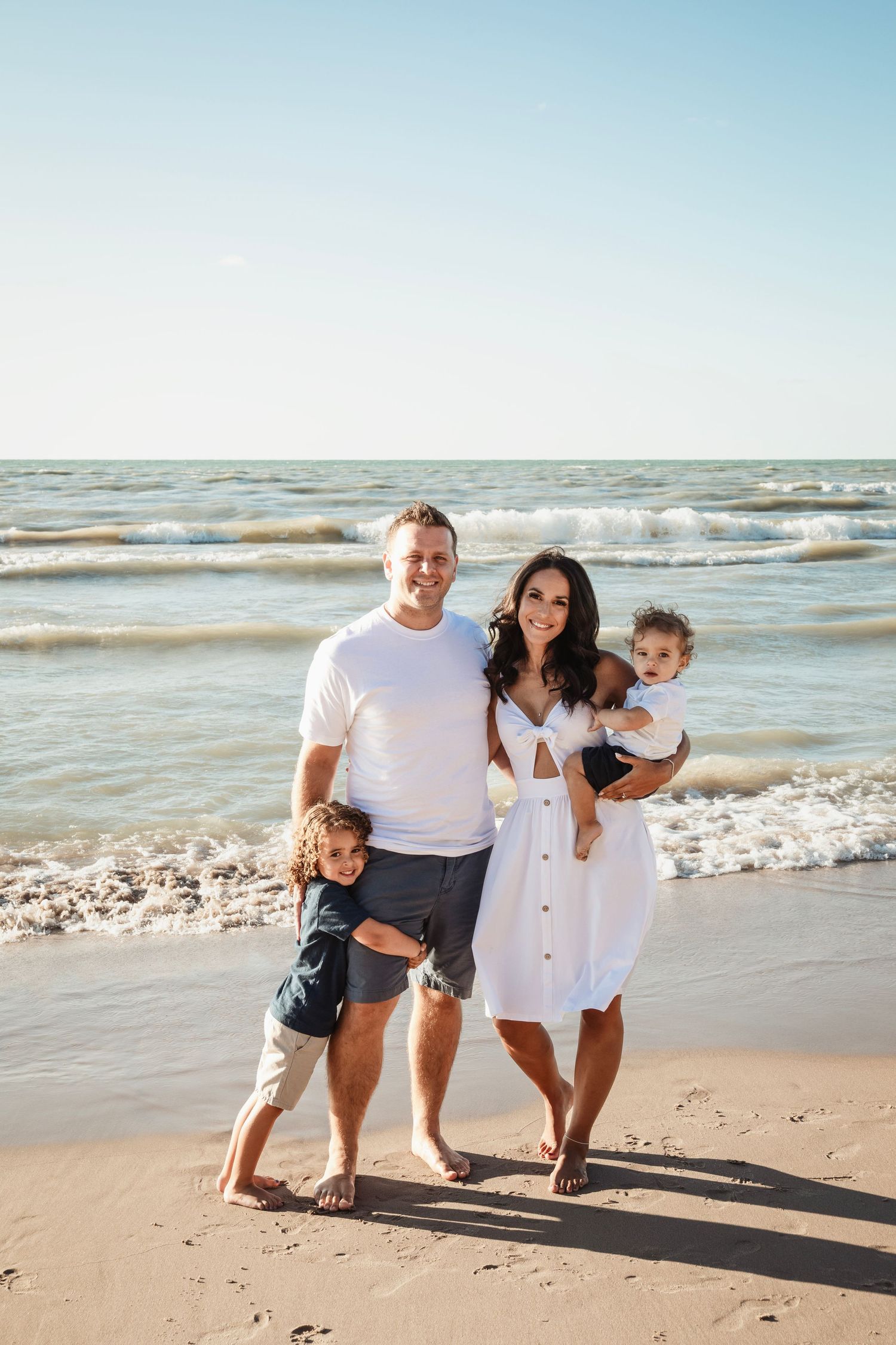 Family of four in white outfits posing together on sunny beach.