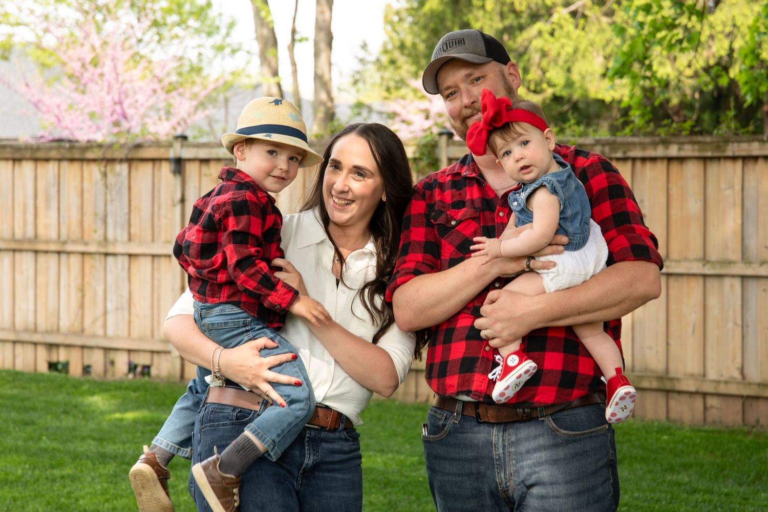 Happy family of four in matching red plaid shirts posing together in a green backyard on a sunny day.