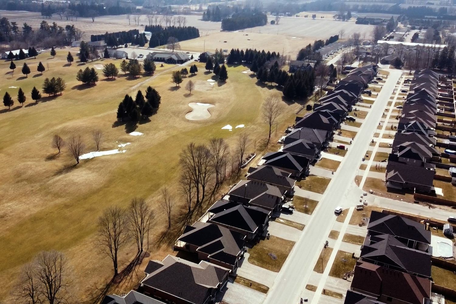 Aerial view of suburban houses along a straight road adjacent to a golf course with winter grass and scattered trees.