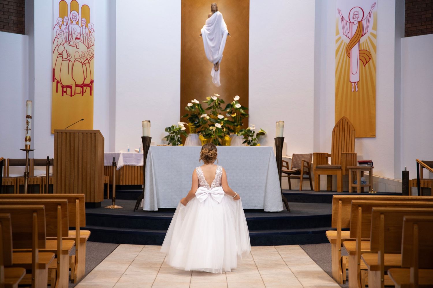 A young girl in a church interior with religious artwork and wooden pews bathed in warm lighting for her first communion celebration.