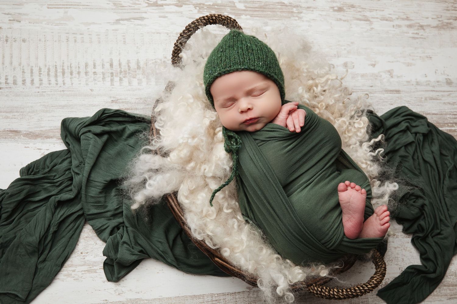 Newborn photography set featuring a dark green swaddle and knit hat on a white wooden background.