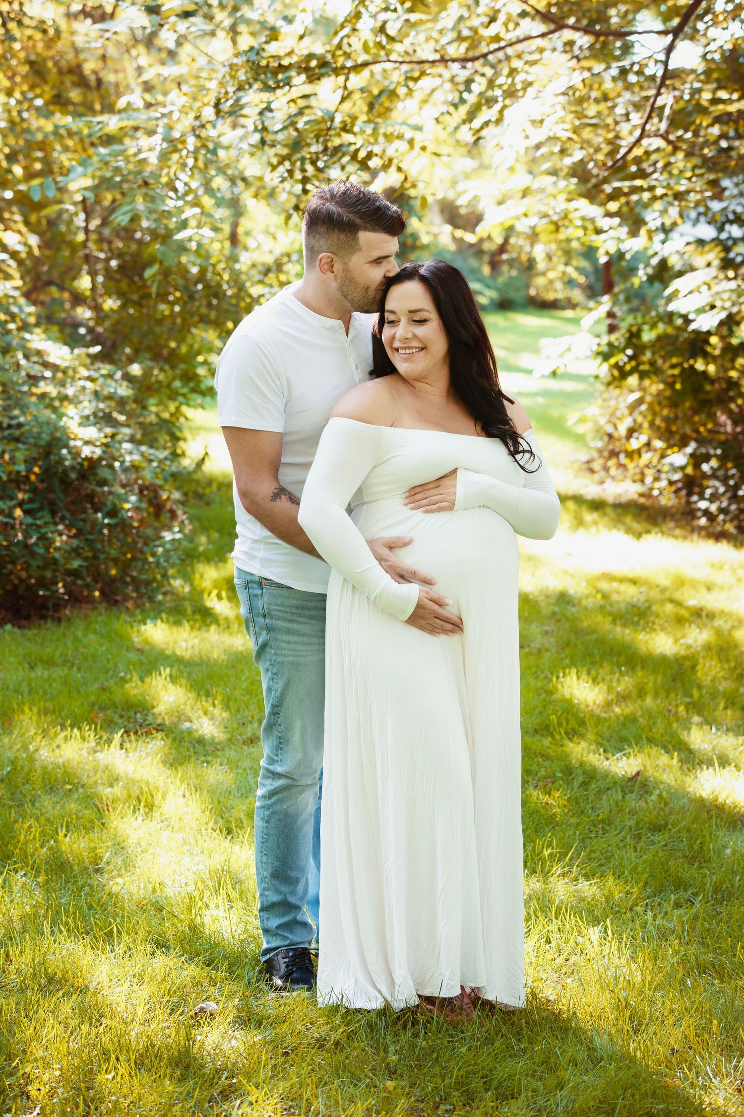 Couple shares intimate moment during maternity photoshoot outdoors in white flowing dress.