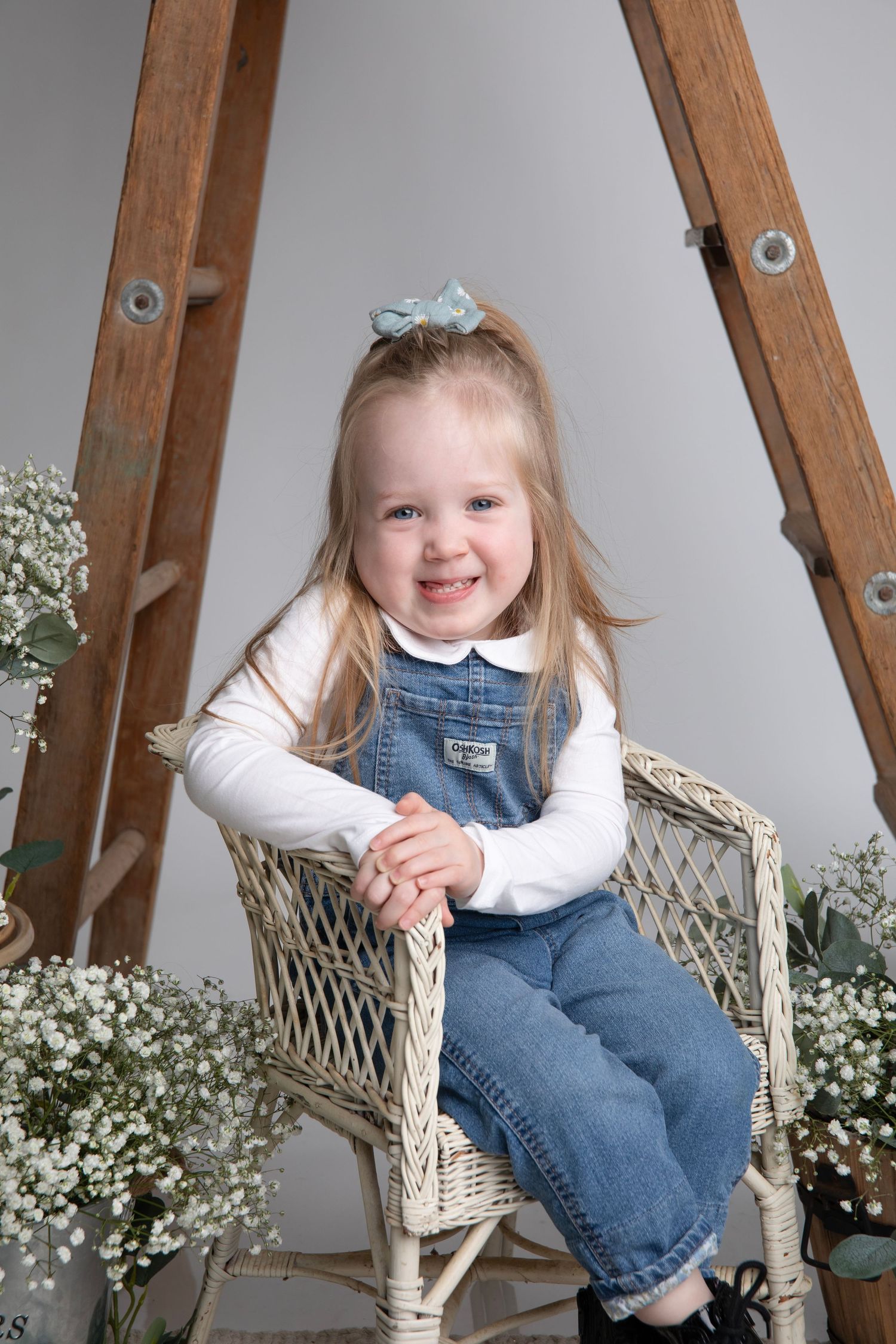A child in denim overalls sits on wicker chair surrounded by baby's breath flowers.