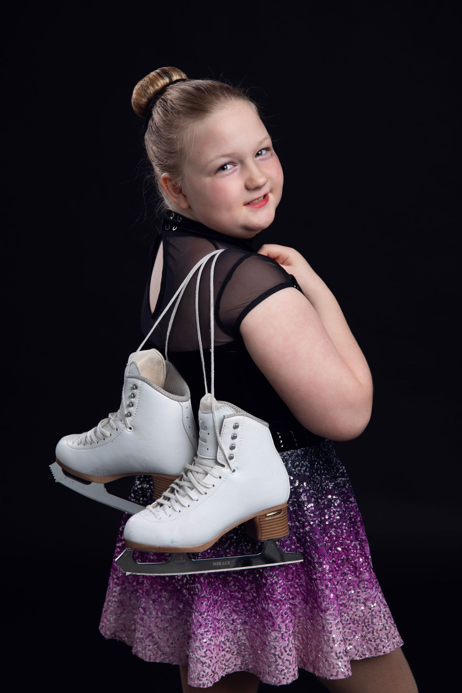 A girl with her figure skates, in a purple gradient dress poses with white ice skates against a dark background.