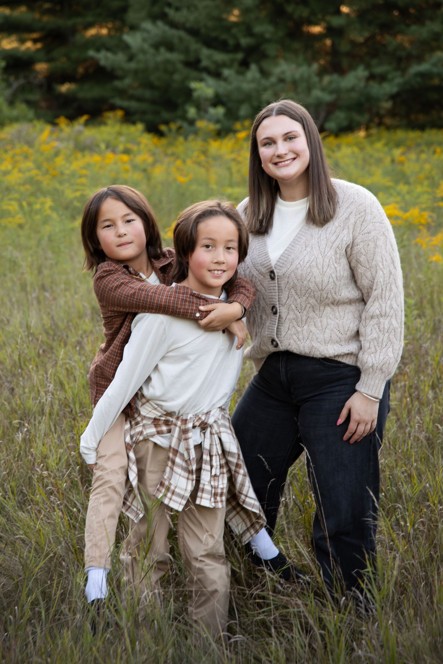 Three friends wearing cozy sweaters pose together in a field of yellow wildflowers on an autumn day.