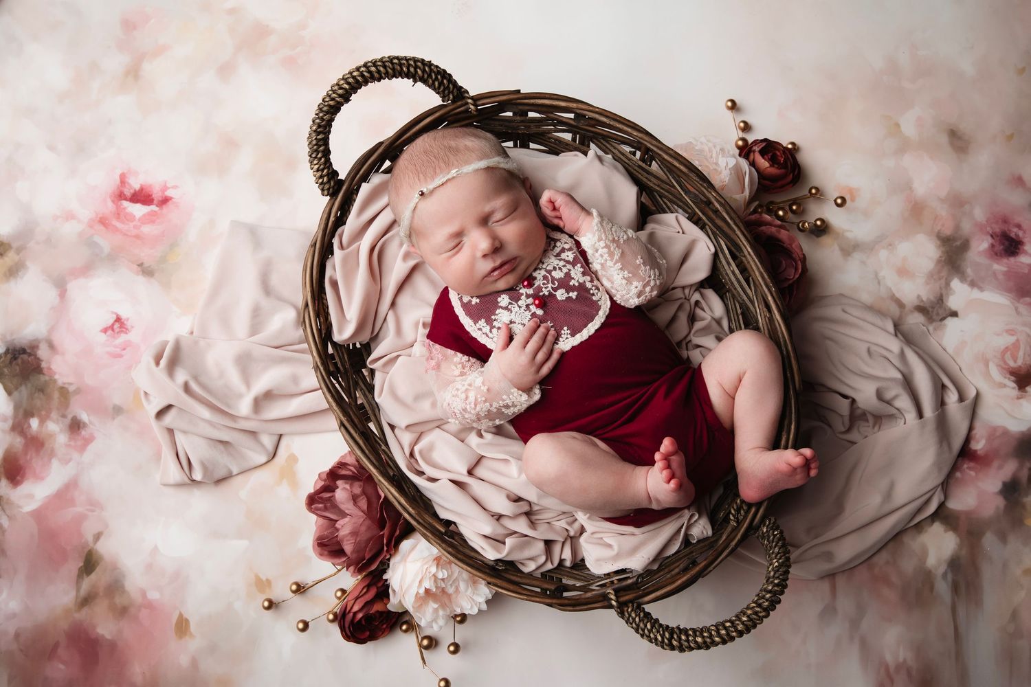 Sleeping newborn in red outfit lies in woven basket surrounded by soft pink rose petals and fabric.