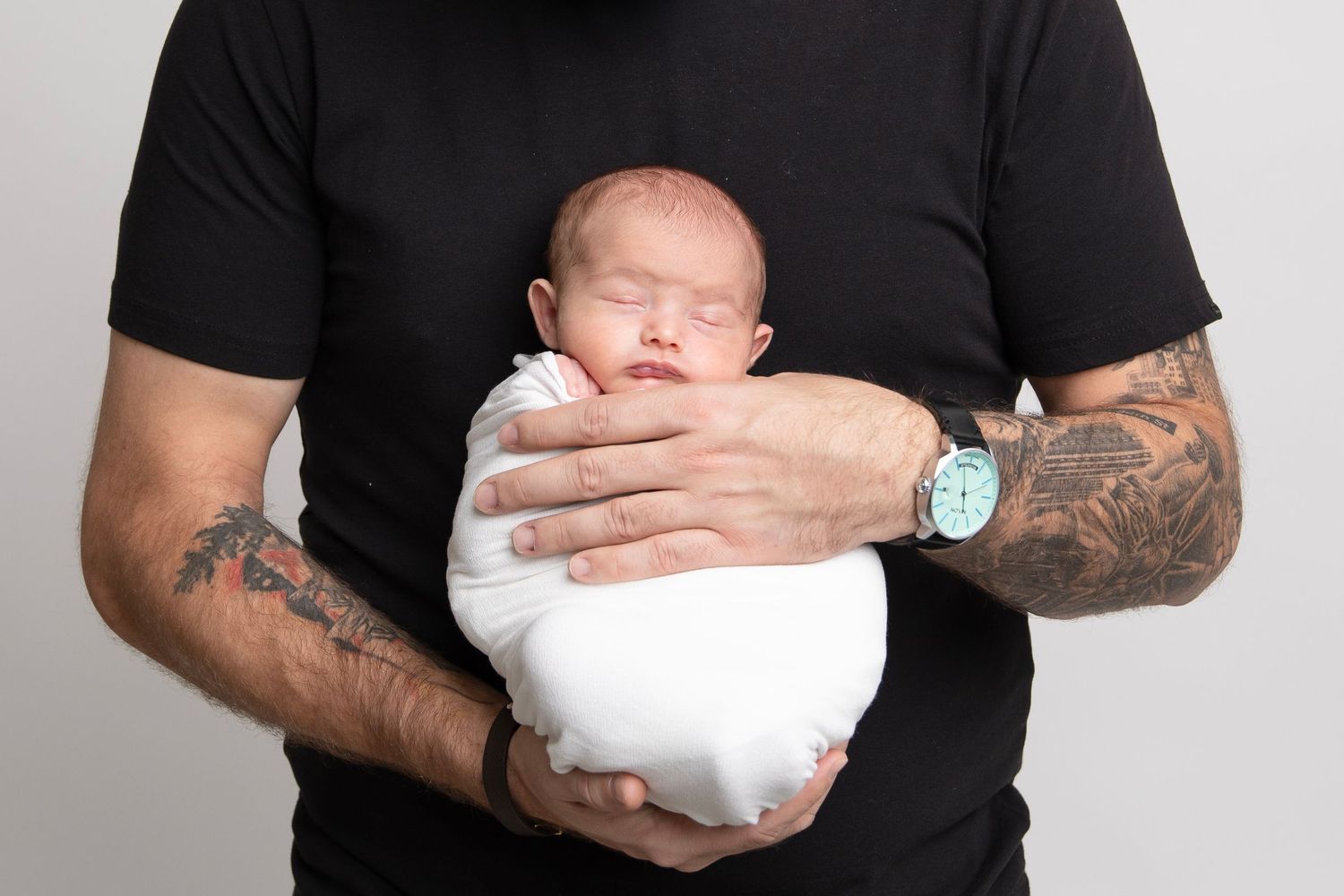 Parent with tattoo sleeves holding sleeping newborn baby swaddled in white.