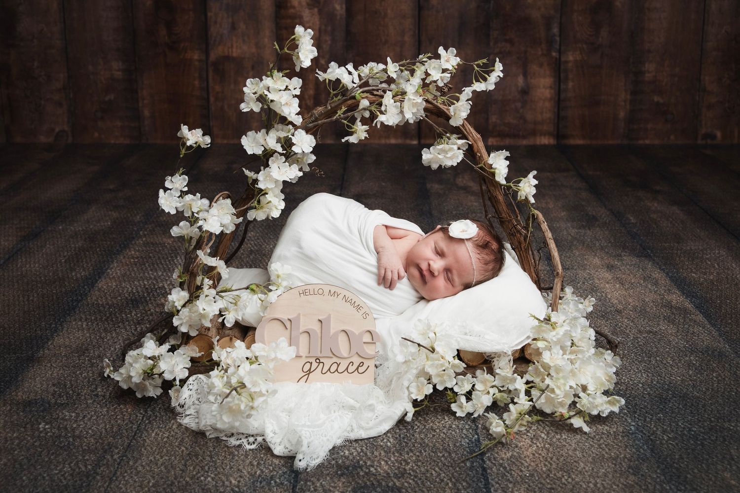 Newborn wrapped in white fabric sleeping beneath white cherry blossom arch on dark backdrop.