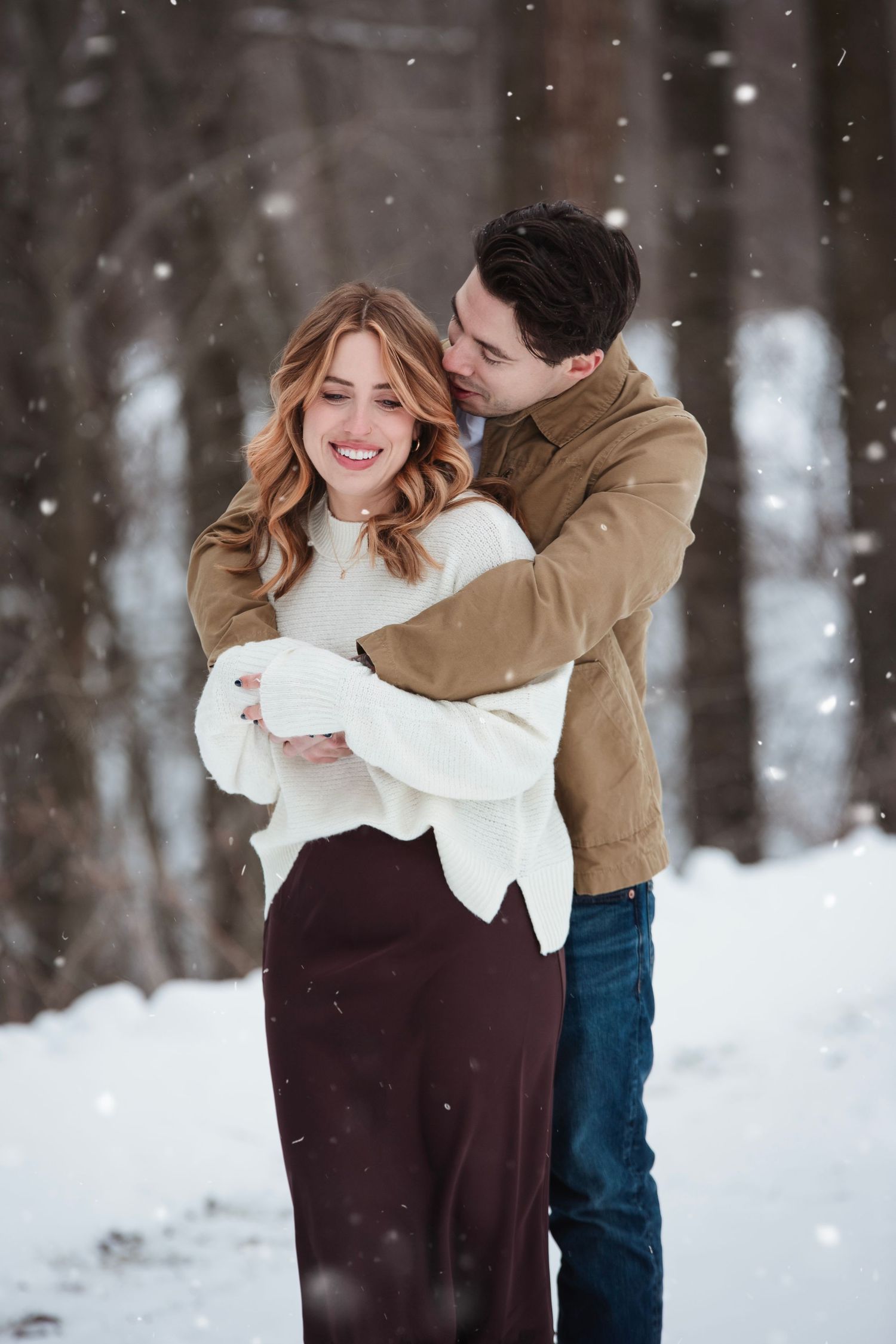 A couple shares a tender moment in a snowy winter forest setting wearing white and brown clothing.