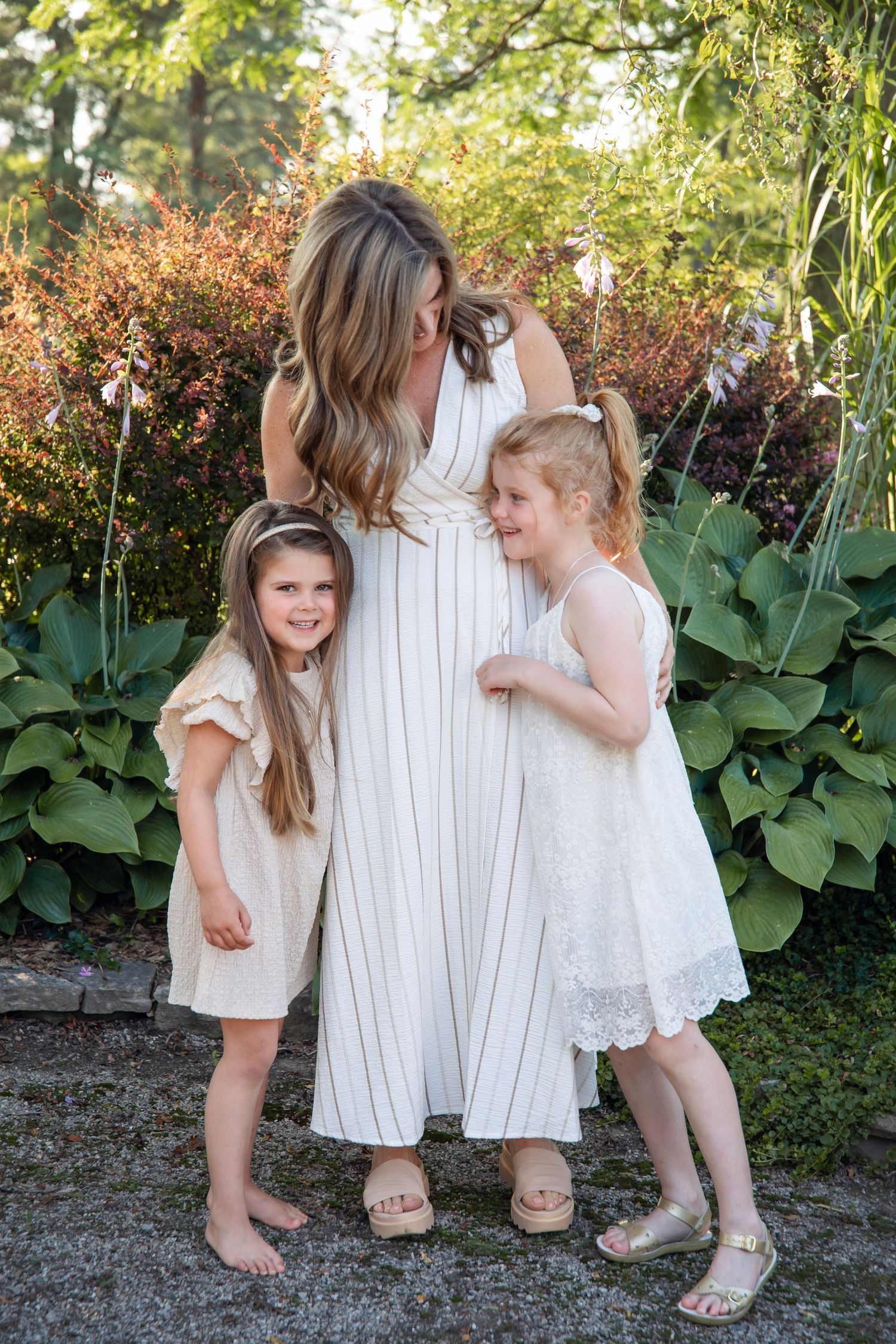 Mother in white striped dress stands with two young daughters in garden setting during summer.
