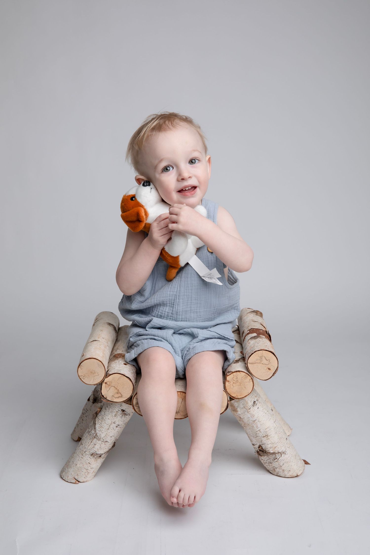 A child in gray overalls sits on a rustic log bench holding a stuffed toy.