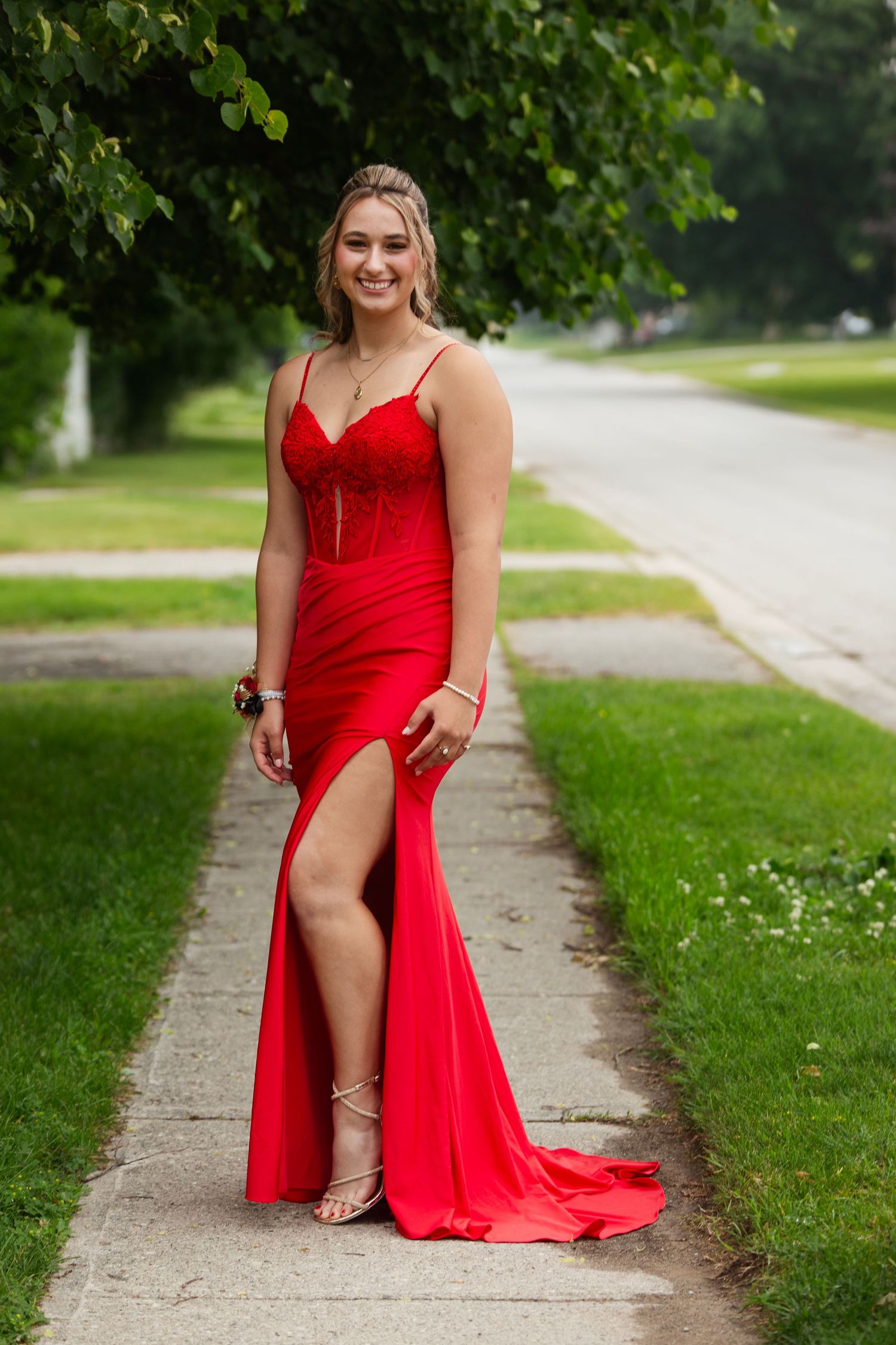 A woman in a stunning red formal dress poses on a tree-lined sidewalk.