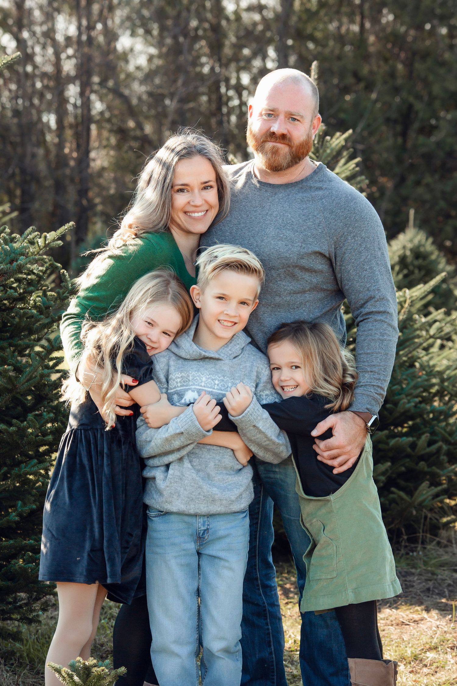 Family of five poses together in front of evergreen trees during outdoor photo session.