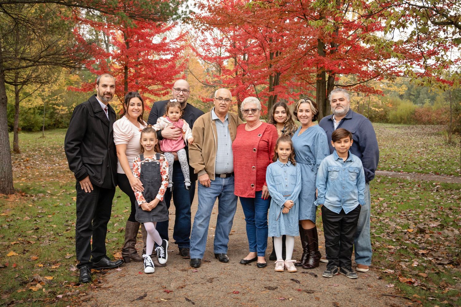 Extended family group portrait outdoors with colorful fall foliage in background.