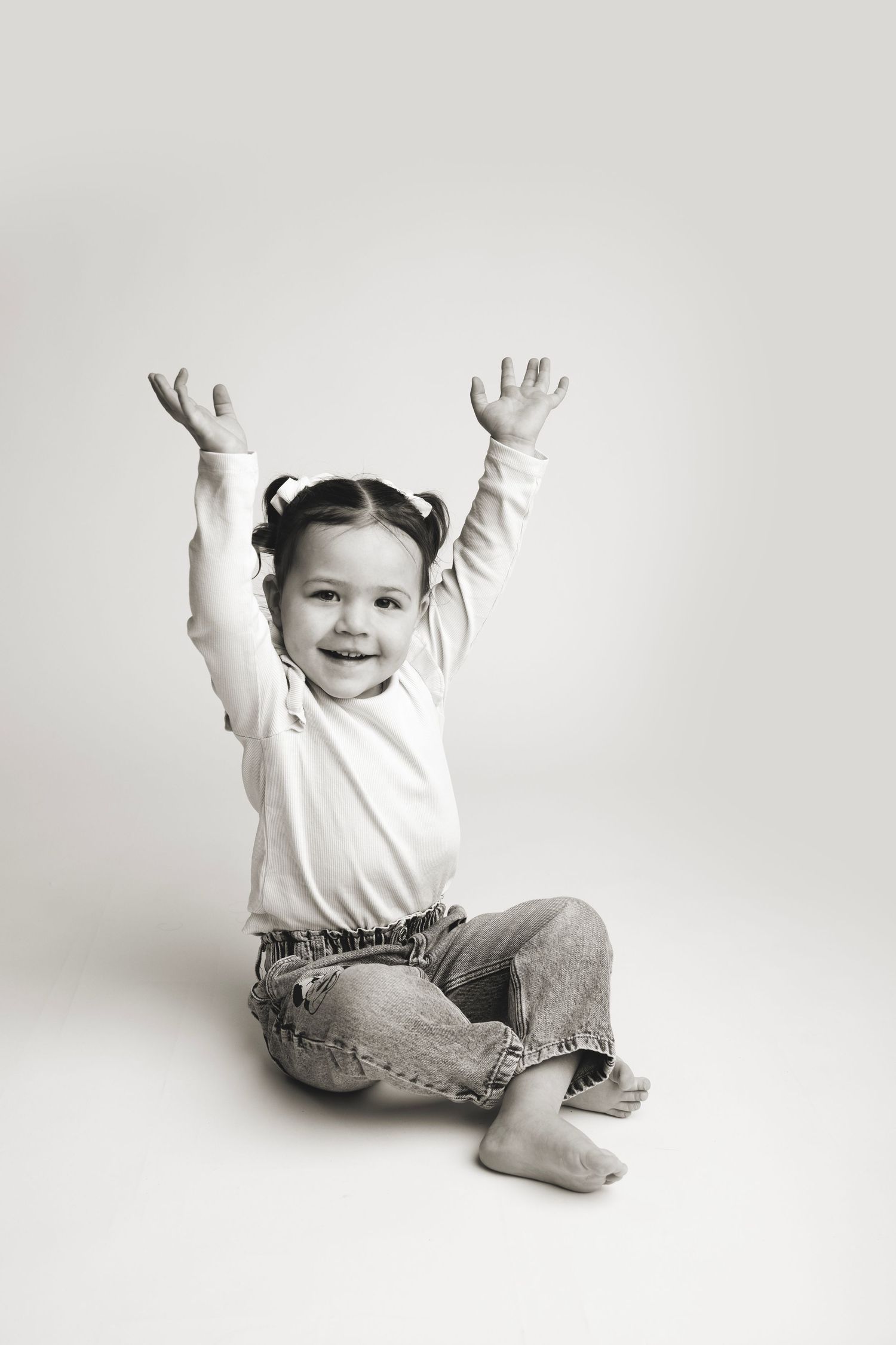 Black and white portrait of a young child sitting on ground wearing jeans and white top with arms raised joyfully.