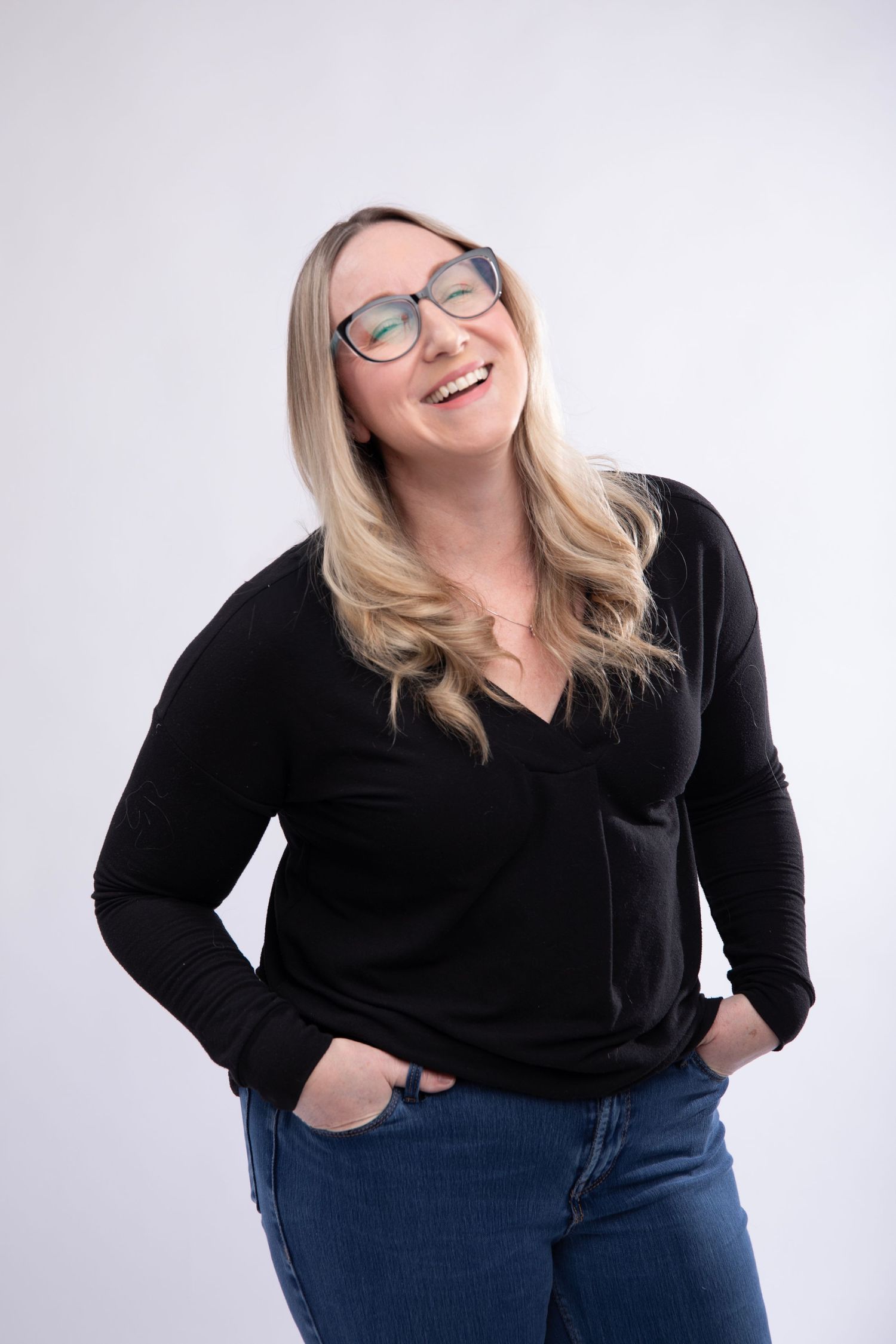 A cheerful person in glasses, black sweater and jeans laughing with hands in pockets against white backdrop.