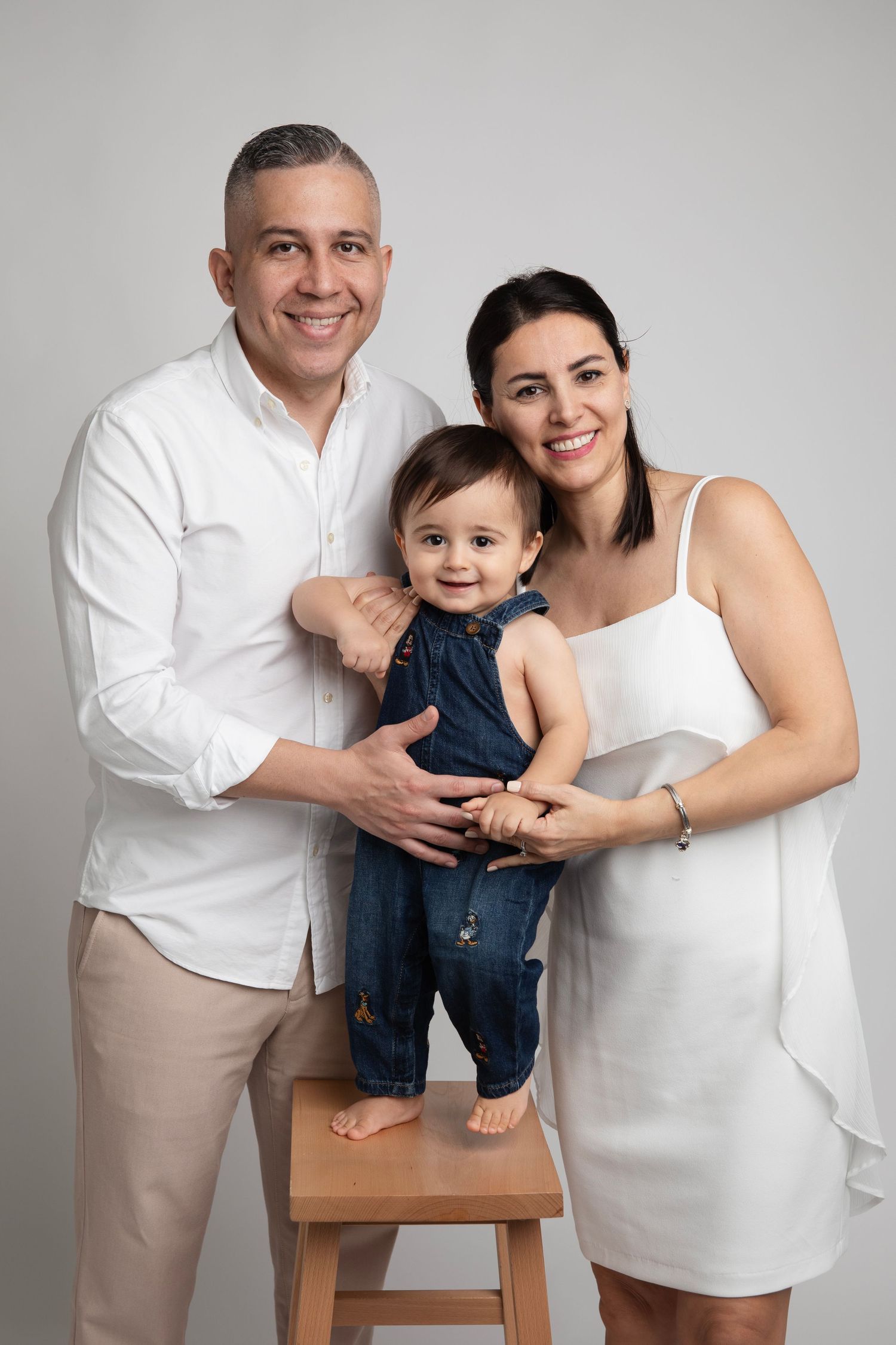 A family wearing white and beige outfits poses together in a series of minimalist studio portraits.