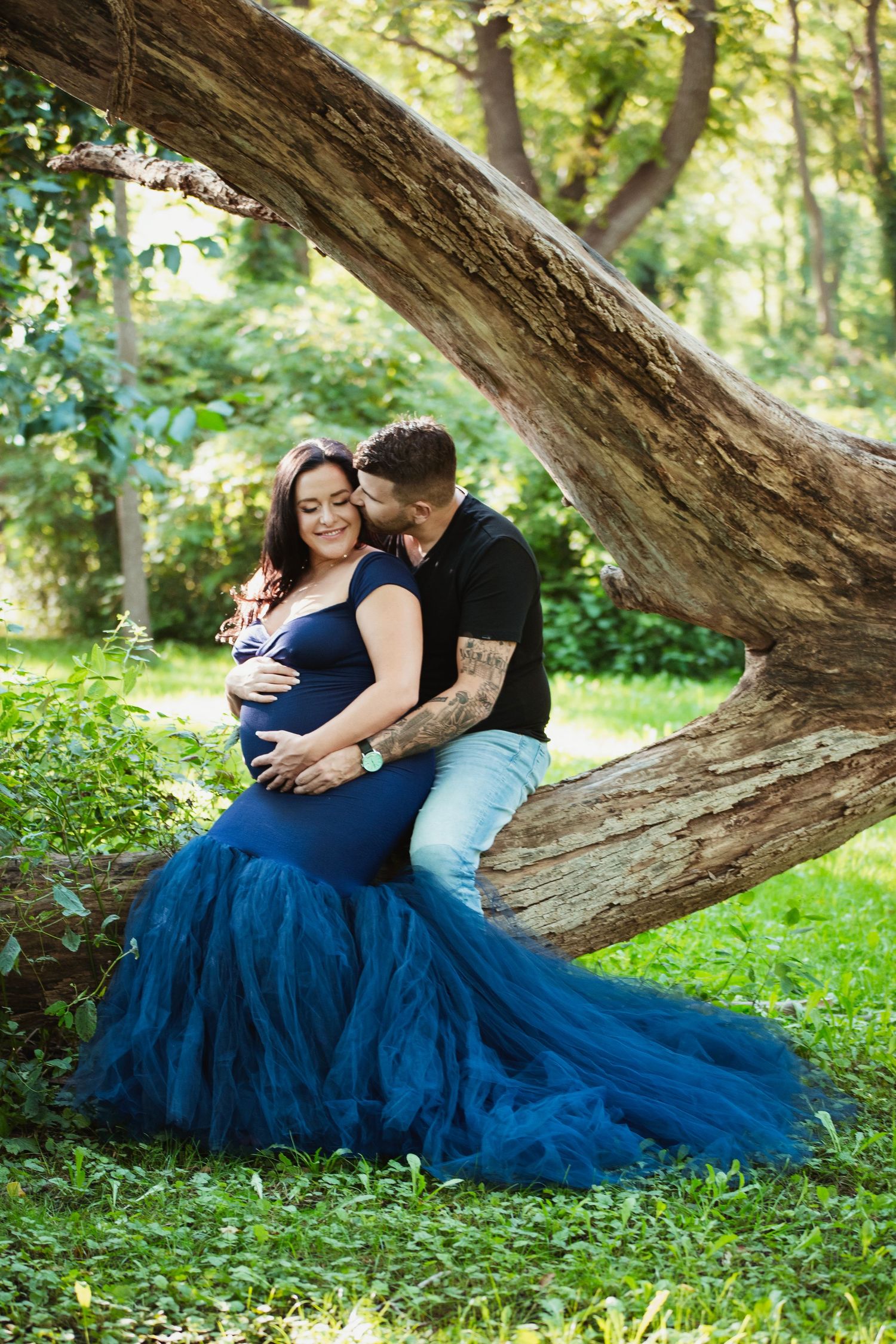 Expecting couple embraces by fallen tree trunk with flowing navy blue tulle gown.