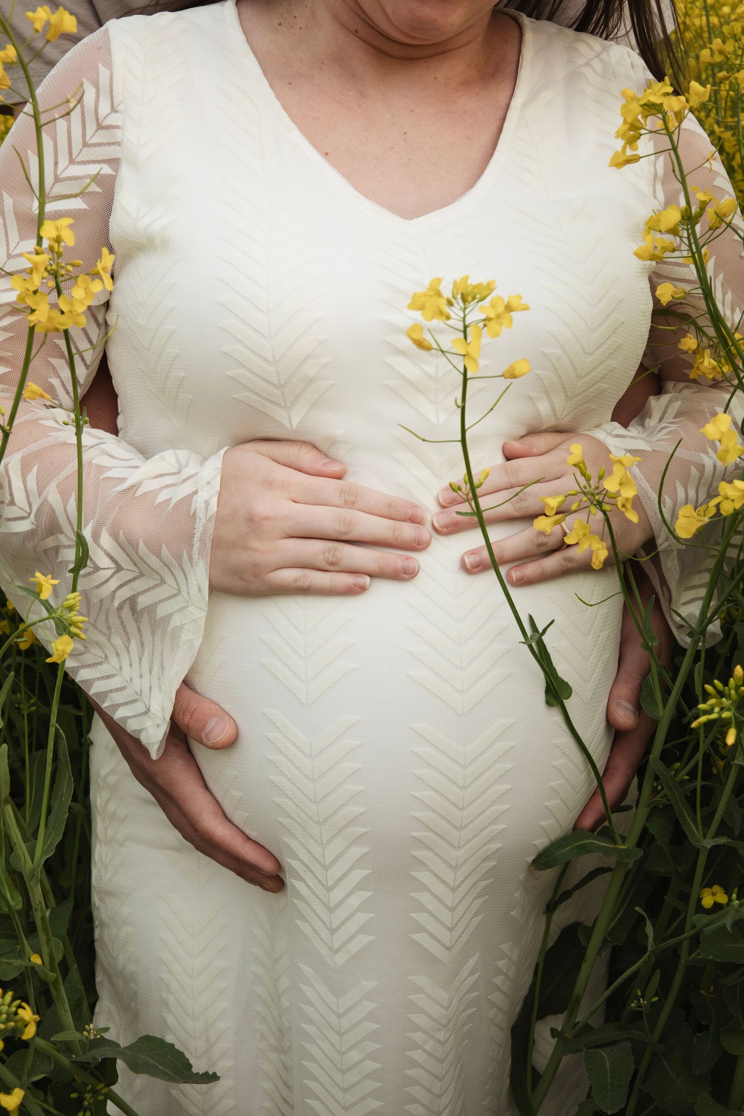 Pregnant woman in white dress holding yellow flowers against a backdrop of blooming forsythia in a garden setting.