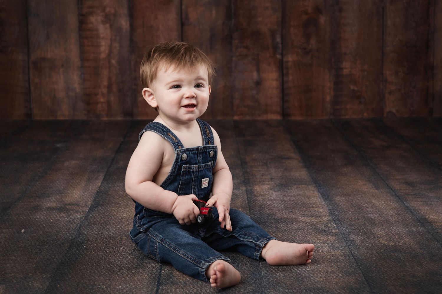 A small toddler in denim overalls sits on a wooden floor with a charming expression.