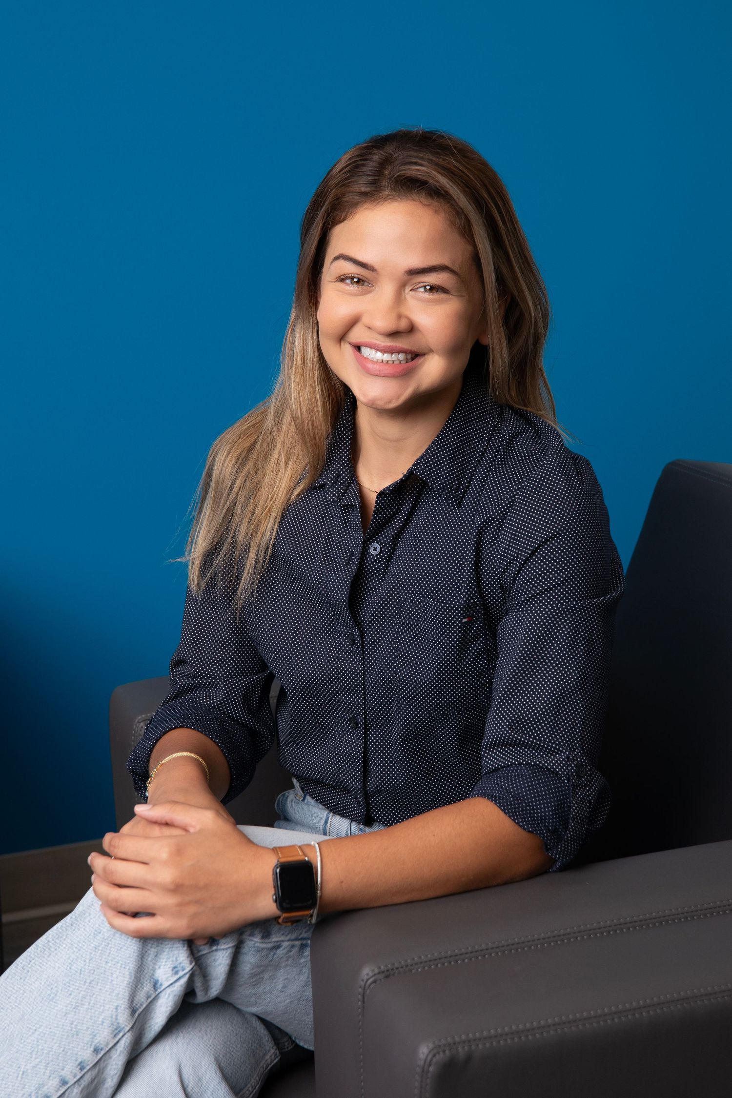 A person in a navy button-down shirt and jeans sits casually against a blue background wearing a smart watch.