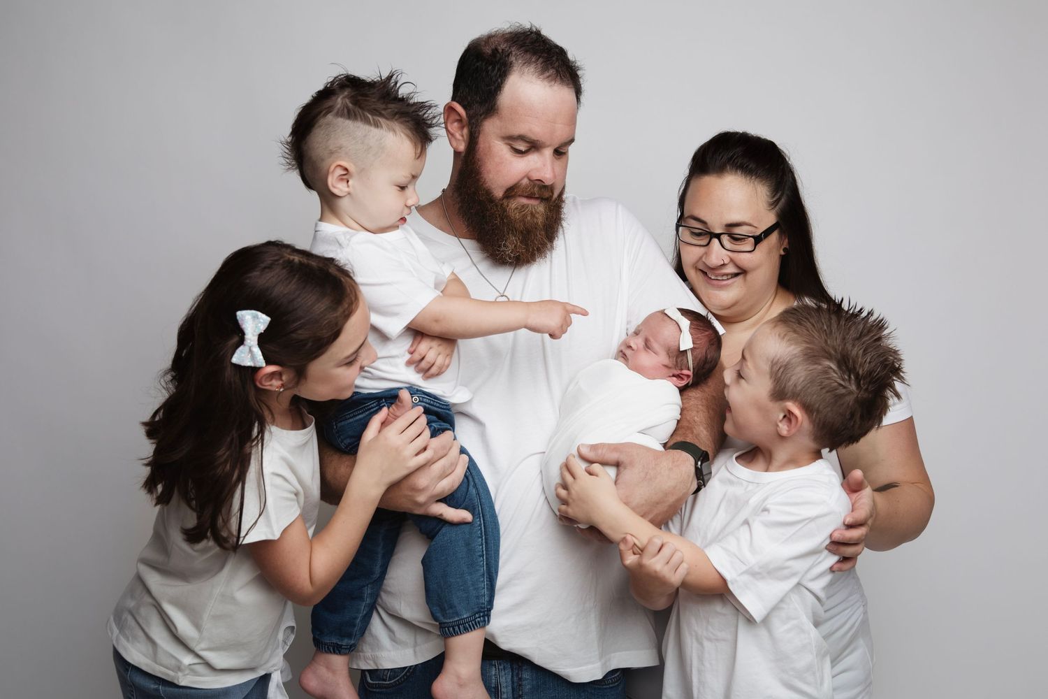 Family portrait with parents and three children interacting joyfully against grey background.