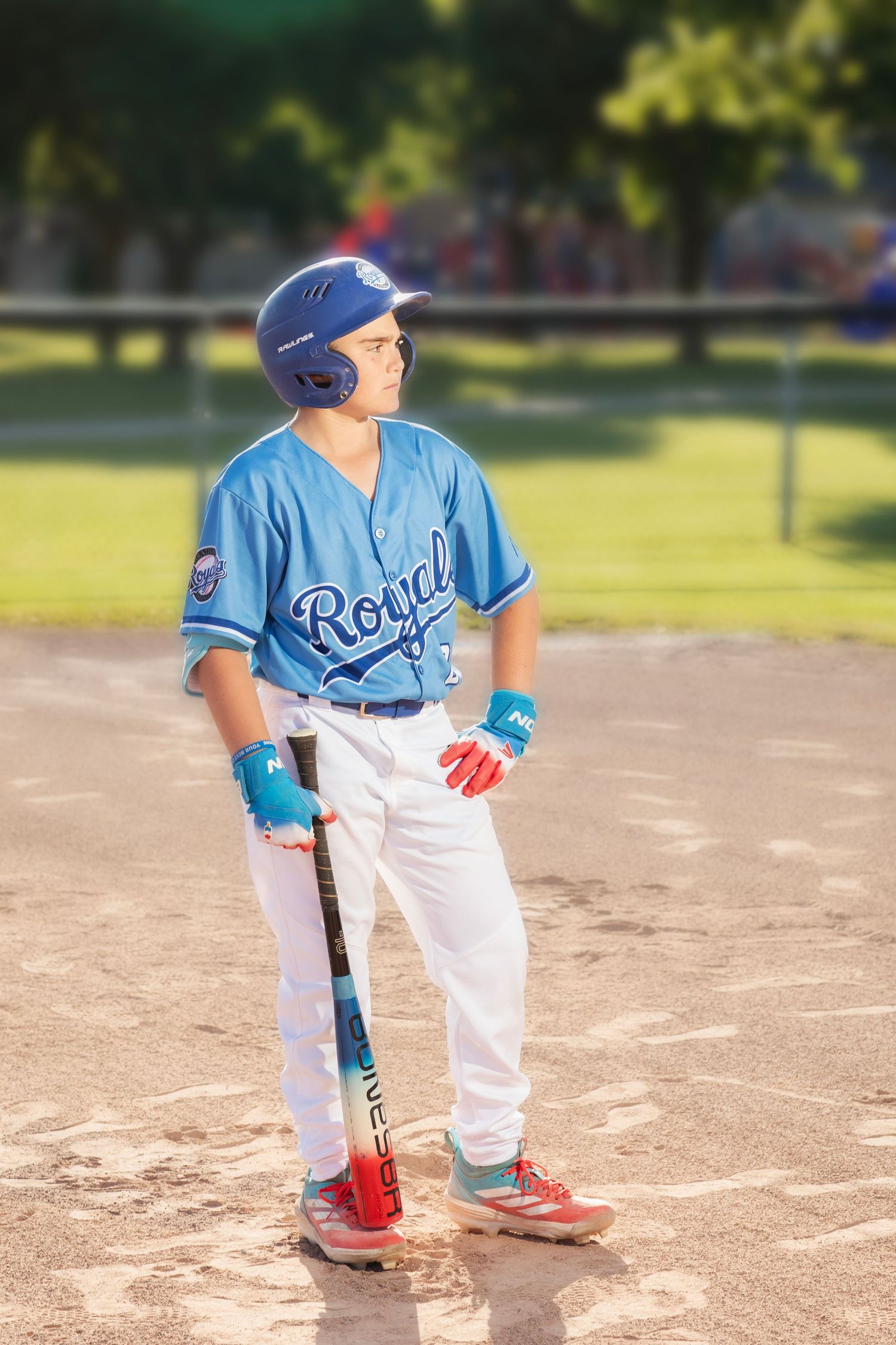 Young baseball player in blue Royals uniform holding bat on dirt field wearing helmet and batting gloves.