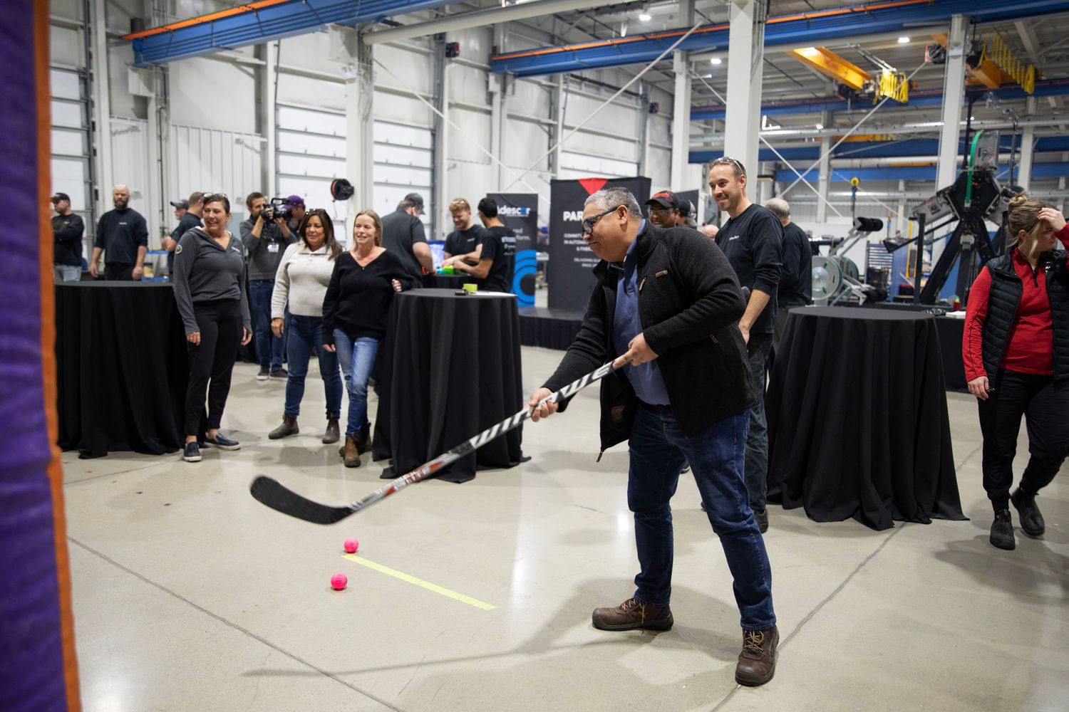 People gather around display tables in an industrial space as someone takes a shot at a hockey game setup.