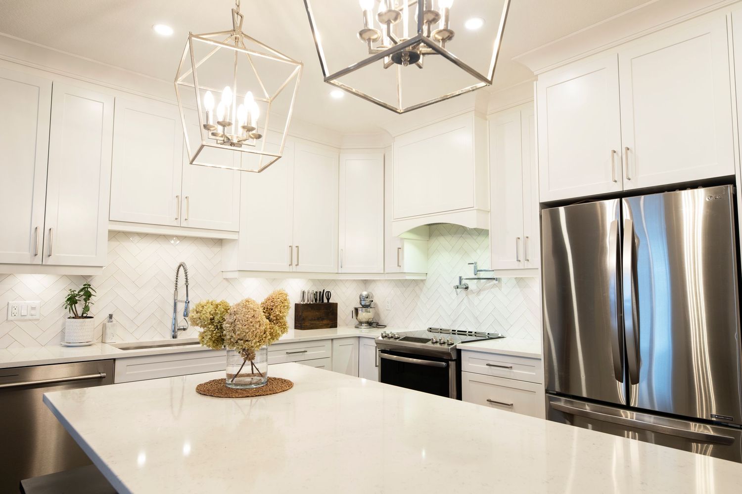 Modern white kitchen with crystal chandeliers, quartz countertops, stainless steel appliances and bright cabinet lighting.