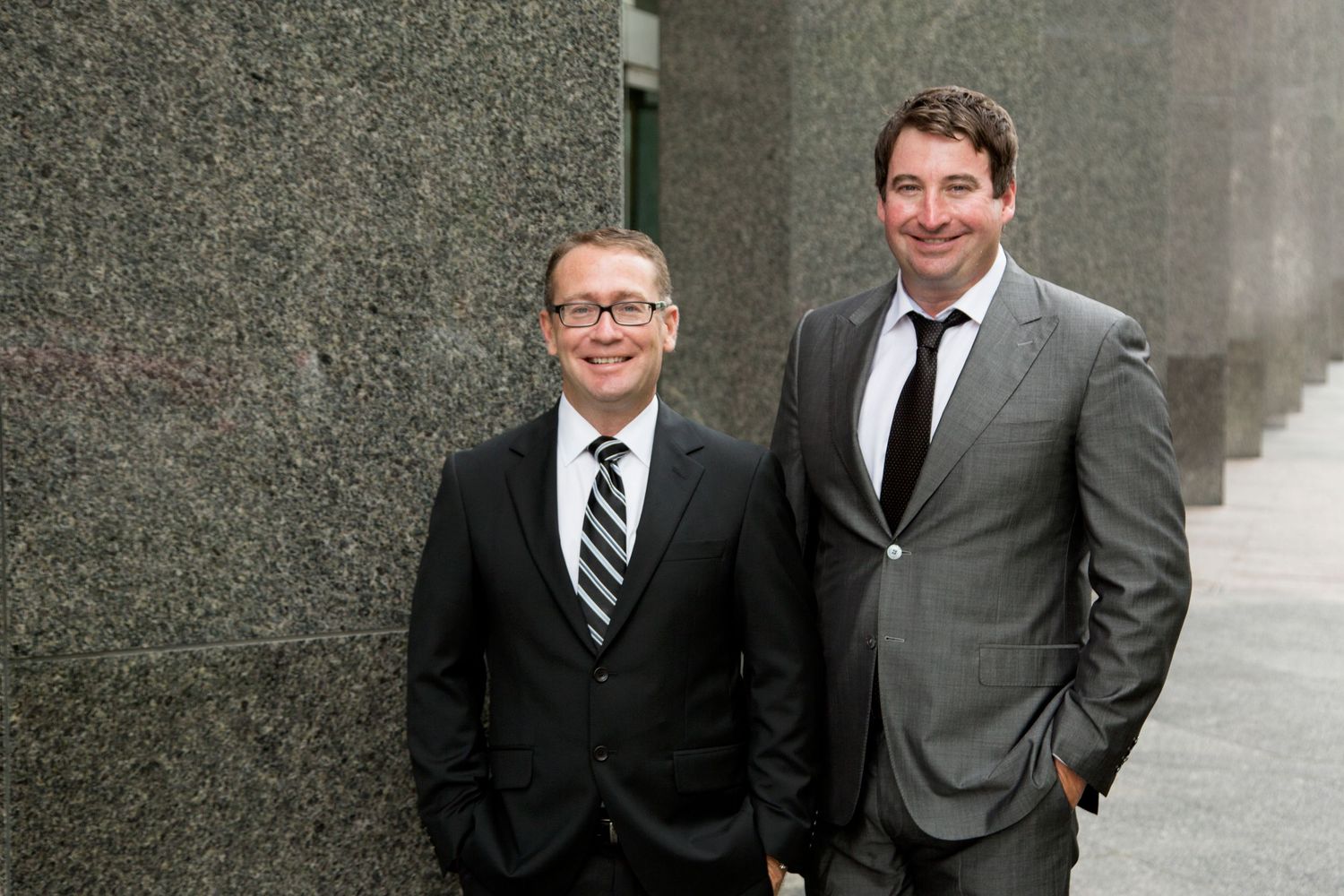Two business professionals in dark suits stand together smiling against a grey wall background.